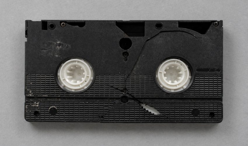 A VHS cassette shell with the bottom panel removed to expose the two metal spool hubs, the kind of physical damage that requires remounting the tape onto a donor shell