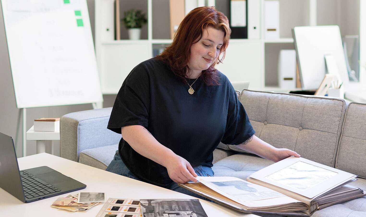 a young woman with red hair and a black shirt works with large images in a portfolio in a studio setting with a laptop to her left and additional photo materials on the table in front of her