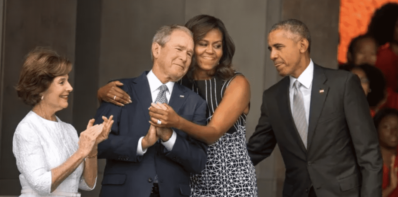 a color image of barbara bush, george w bush, michelle obama and barack obama, standing outside the funeral of george hw bush. michelle obama is hugging president bush while all four subjects smile