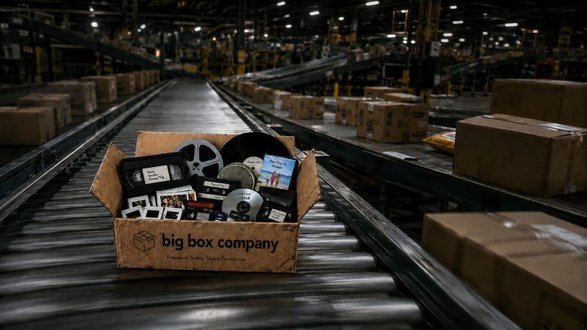 A worn open box of family tapes, film reels, photos and slides moving down a conveyor belt in a dark parcel sorting warehouse