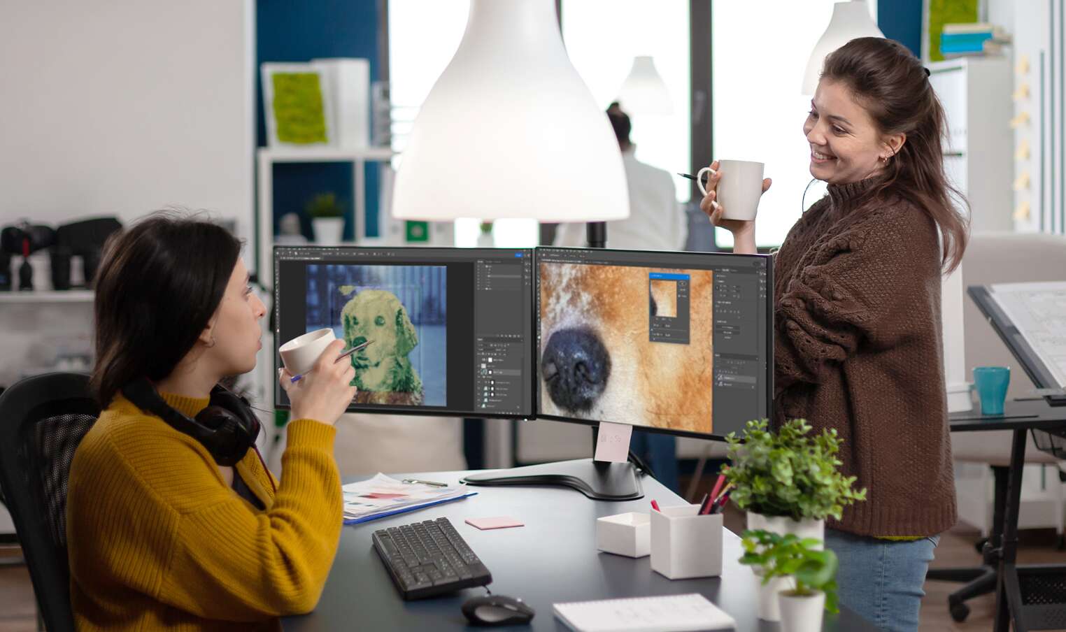 two women converse with a computer desk between them. both are holding coffee mugs. one of the women is sitting, and appears to be working on a photo restoration project on two computer monitors, while the other smiles at her.